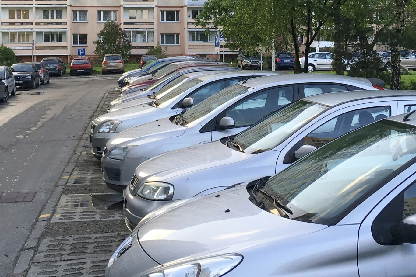 Często wyjeżdżając z parkingu nie zauważamy kartki umieszczonej na tylnej szybie. Lepiej zachować czujność, bo to może być początek naszych problemów. Szereg srebrnych i szarych samochodów osobowych zaparkowanych obok siebie na parkingu osiedlowym, w tle bloki mieszkalne i drzewa po obu stronach ulicy.