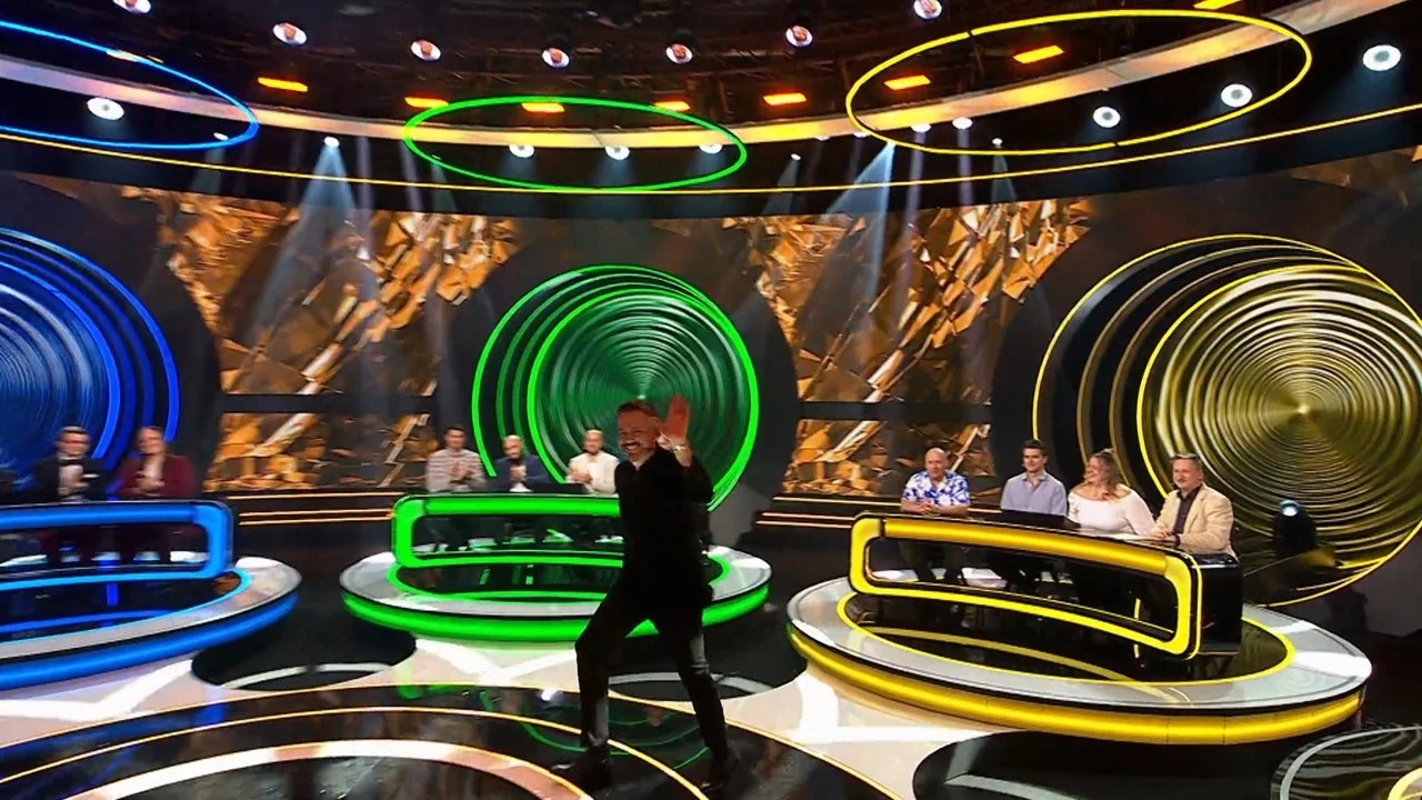 Krzysztof Ibisz jak zawsze w formie. Awatura o kasę rozpoczęta Uczestnicy programu telewizyjnego siedzą przy okrągłych stołach z kolorowymi podświetleniami, w tle widoczne są okręgi w różnych barwach, na pierwszym planie osoba prowadząca w ruchu.