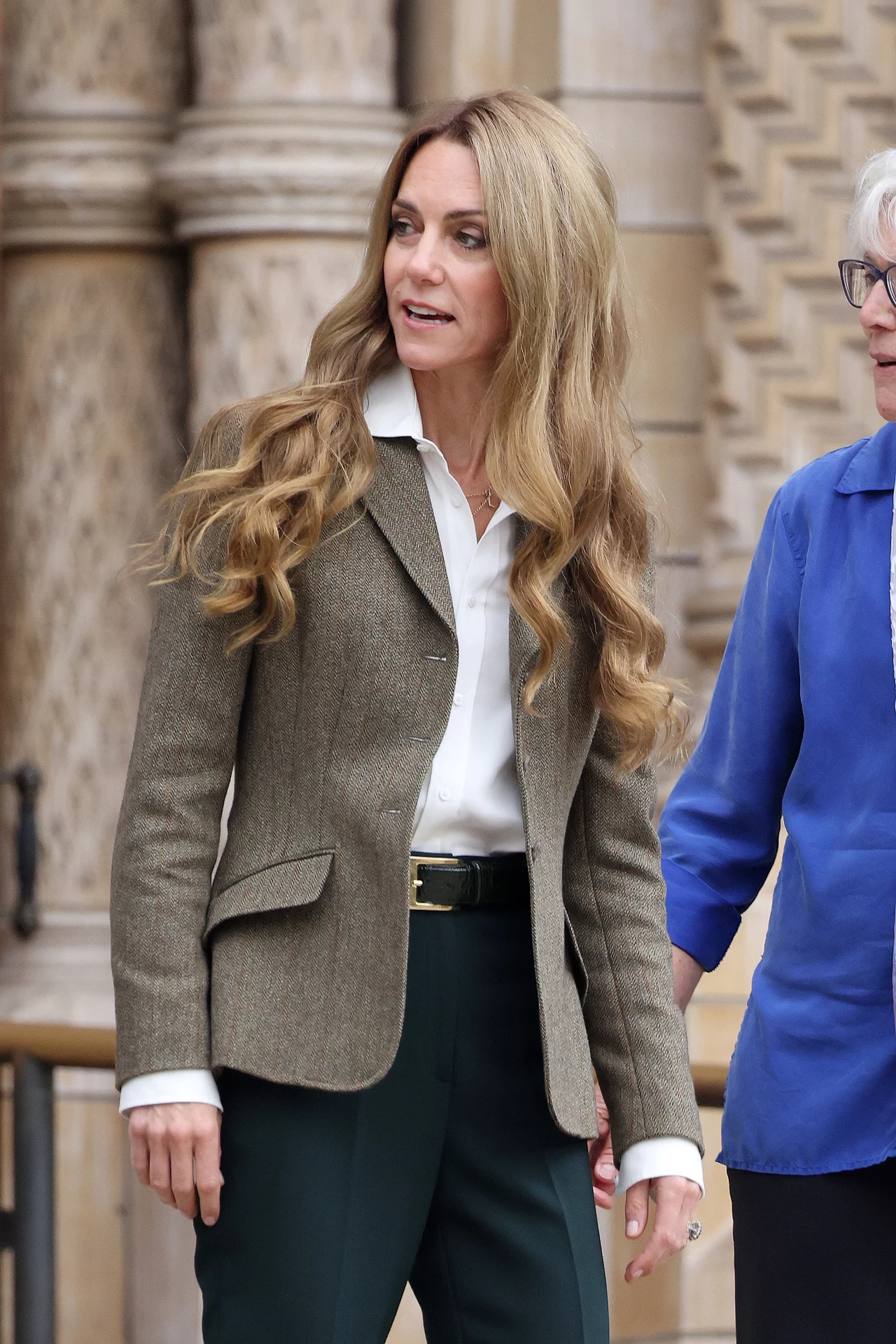 Tak jasnych włosów jeszcze nie widziano u księżnej Kate Kobieta z długimi falowanymi włosami ubrana w elegancką marynarkę i białą koszulę stoi na tle rzeźbionych kamiennych kolumn, obok niej znajduje się druga osoba ubrana w niebieską koszulę, której twarz nie jest w pełni widoczna.