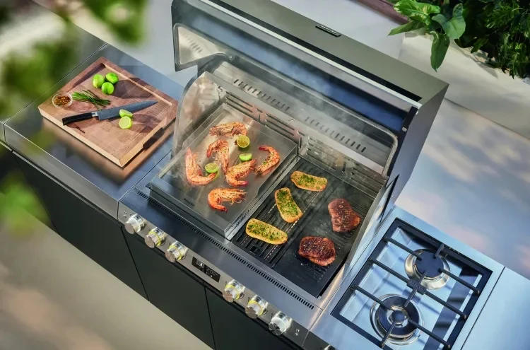 Miele Dreams to inteligentny grill z funkcją Fire Pro IQ. Grill gazowy z rusztem, na którym pieką się steki oraz kawałki kurczaka z limonką, po lewej stronie deska z pokrojonymi limonkami i nożem; wokół elementy nowoczesnej kuchni z widocznym palnikiem gazowym.