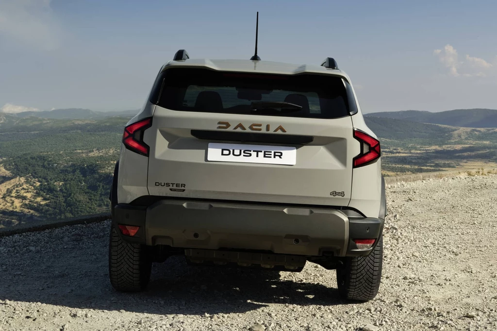 W zależności od modelu średnie zużycie benzyny będzie wynosić 5,5-5,6 l/100 km, a LPG 7,1-7,2 l/100 km. SUV Dacia Duster zaparkowany na szutrowej drodze, z widokiem na rozległe, pagórkowate tereny i góry w tle, widoczny od tyłu z wyraźnym emblematem marki.