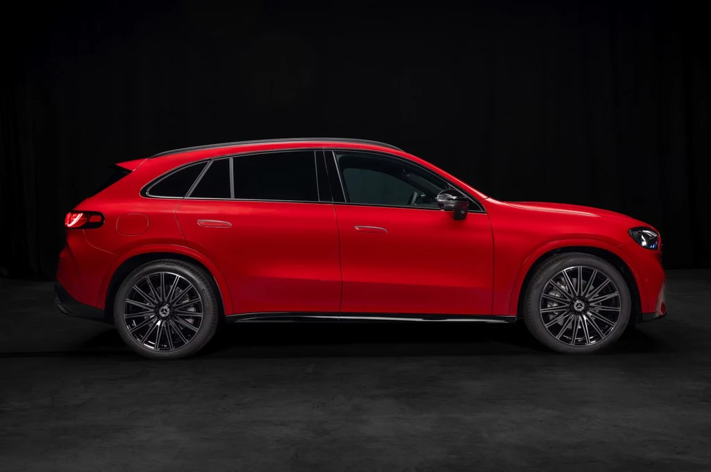 Powiększony rozstaw osi i króciutkie zwisy – to zapowiedź naprawdę przestronnej kabiny Czerwony SUV Mercedes-Benz z widocznymi czarnymi felgami stoi na tle ciemnego tła, wyraźnie podkreślony aerodynamiczny kształt nadwozia oraz przyciemniane szyby.