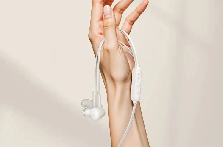 Słuchawki douszne na kablu zwisające z dłoni uniesionej pionowo na tle jasnego, neutralnego tła.