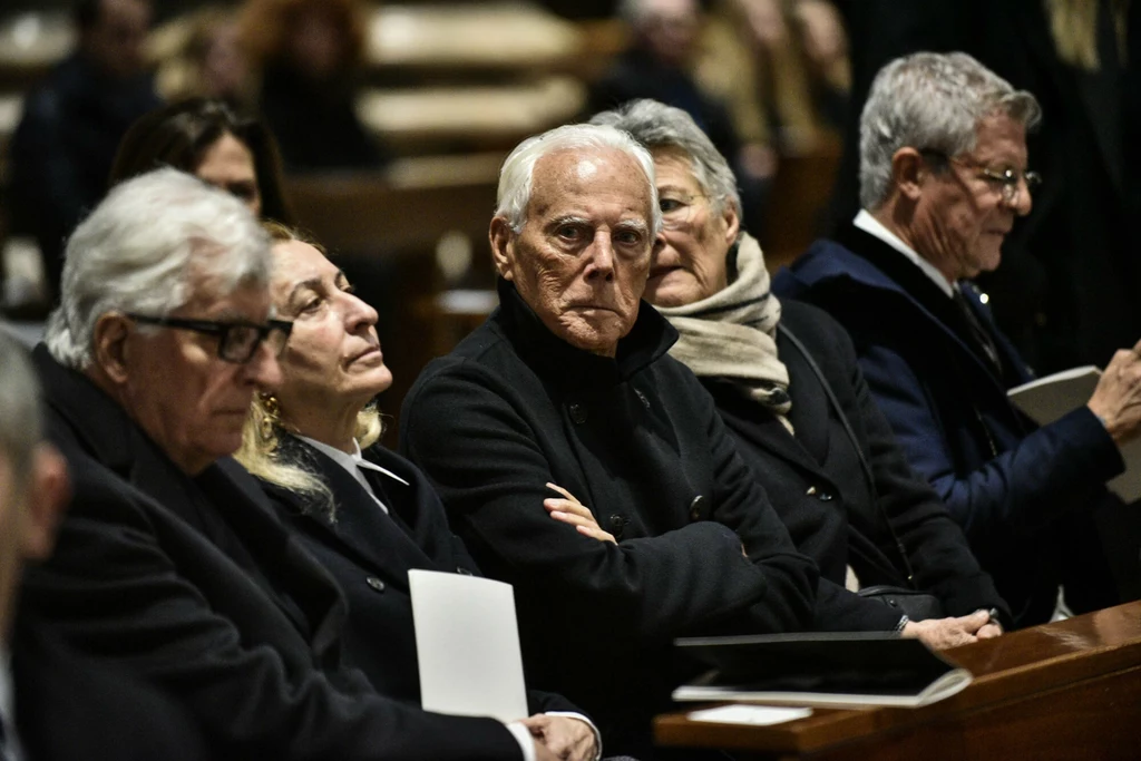 Grupa starszych, elegancko ubranych osób siedzi w ławkach, prawdopodobnie podczas uroczystości o poważnym charakterze, na twarzach widoczne skupienie i zaduma.