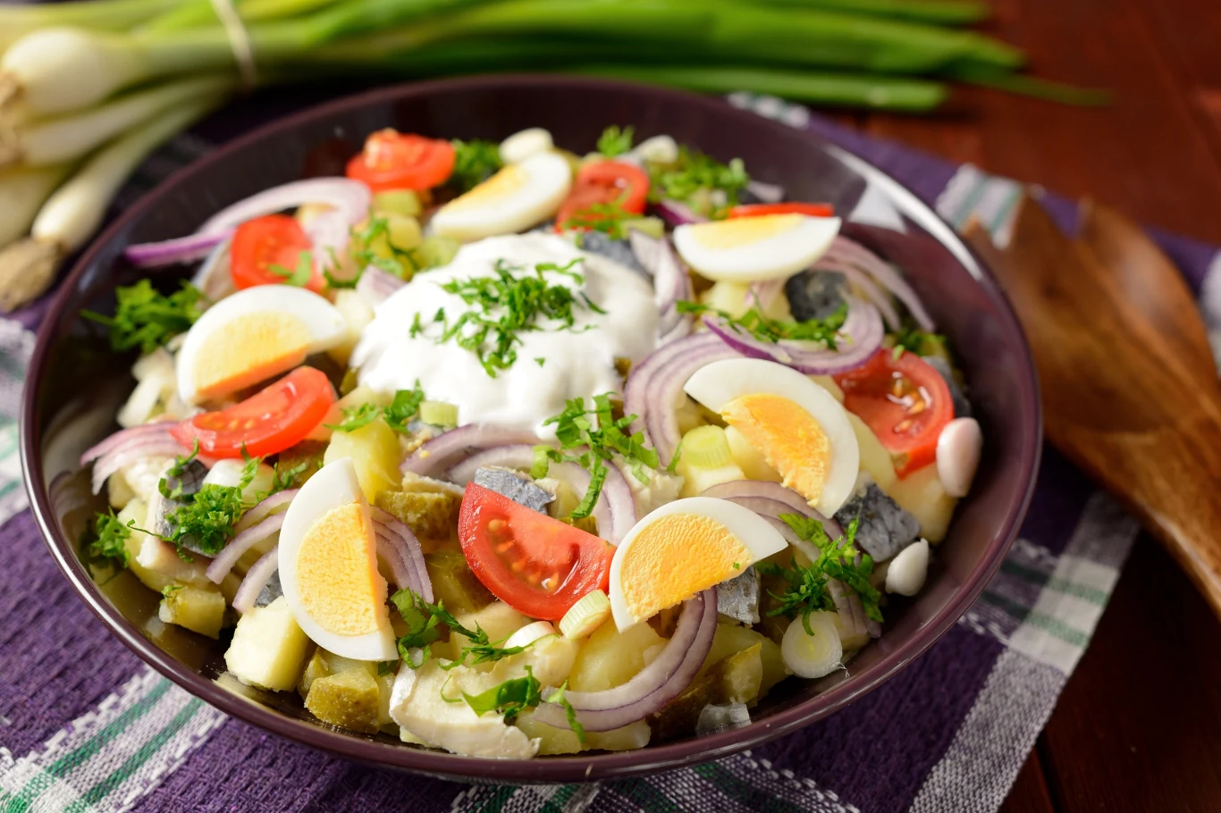 Kolorowa sałatka śledziowa z jajkiem, pomidorem, cebulą, ogórkiem i zielonym koperkiem, udekorowana kleksem śmietany, podana w misce na płóciennym obrusie, obok widoczne świeże szczypiorki i drewniane sztućce.