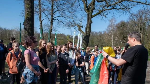 Grupa młodzieży wraz z opiekunem zgromadzona na świeżym powietrzu w parku podczas słonecznego dnia, mężczyzna prezentuje kolorowe flagi lub elementy materiału, dzieci skupione i zainteresowane tematem.