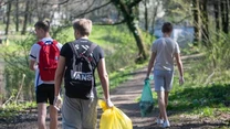 Trzech młodych chłopców niesie worki z zebranymi śmieciami leśną ścieżką, każdy ma założone rękawiczki ochronne, a otoczenie stanowi zielony las i nasłoneczniona ścieżka