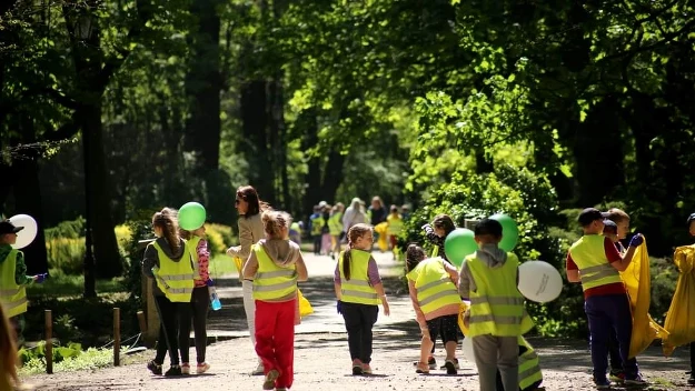Grupa dzieci ubranych w odblaskowe kamizelki sprząta park, trzymając worki na śmieci i balony, wokół nich zielone drzewa i promienie słoneczne oświetlające alejkę.