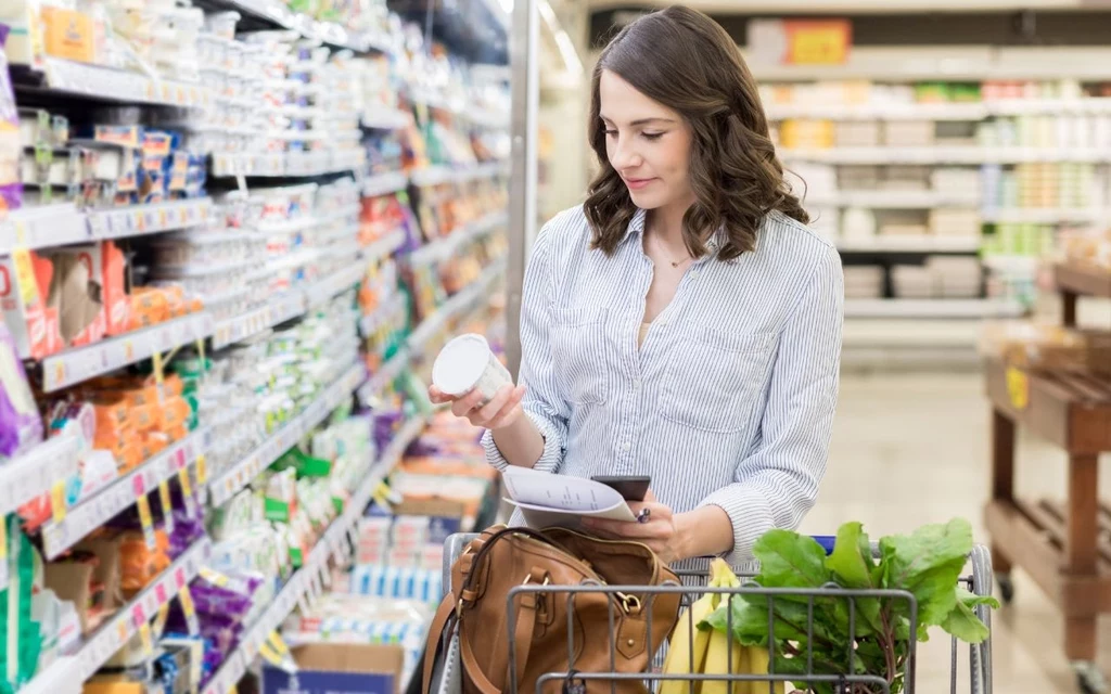 Zawsze sprawdzaj datę ważności produktu w sklepie przed zakupem Kobieta z listą zakupów w ręku stoi przy półce sklepowej, trzymając produkt i analizując jego etykietę, w wózku znajdują się świeże warzywa.
