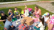 Grupa dzieci siedzi na kocu w ogrodzie, wokół piknikowego kosza, dzieci jedzą przekąski i rozmawiają, w towarzystwie opiekunki, atmosfera jest radosna i beztroska.