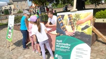 Kilka dzieci i dorosły rozmawiają przy stoliku na świeżym powietrzu podczas eventu ekologicznego, obok widoczne są plansze informacyjne promujące dbanie o planetę oraz ekologiczne inicjatywy.