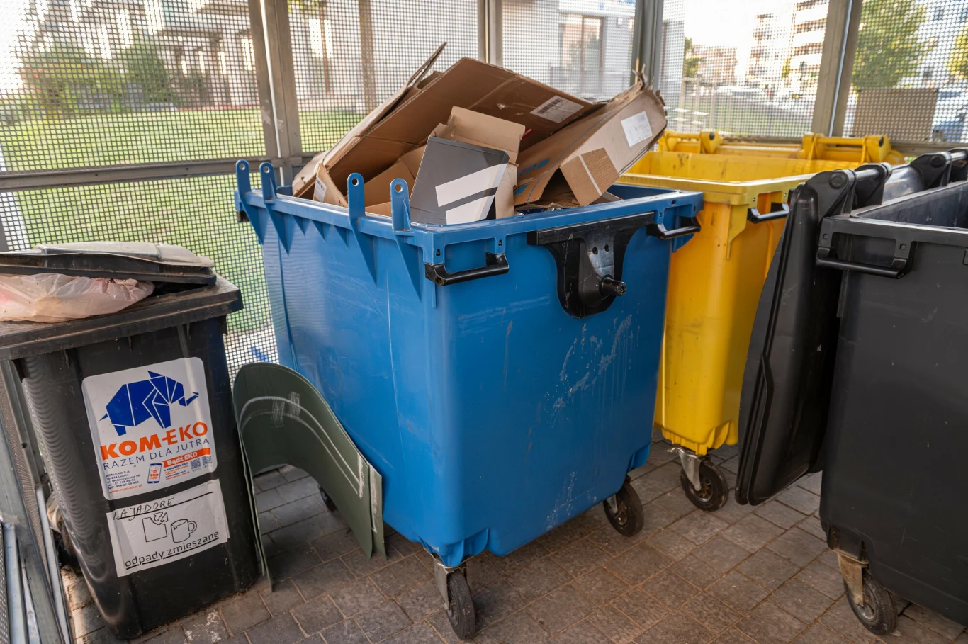 Paragony często błędnie trafiają do niebieskiego pojemnika na odpady. Kontener na odpady z niebieskim pojemnikiem wypełnionym kartonami i innymi odpadami, otoczony przez żółty i czarny pojemnik na śmieci, stojące w wydzielonej przestrzeni w pobliżu budynków mieszkalnych.