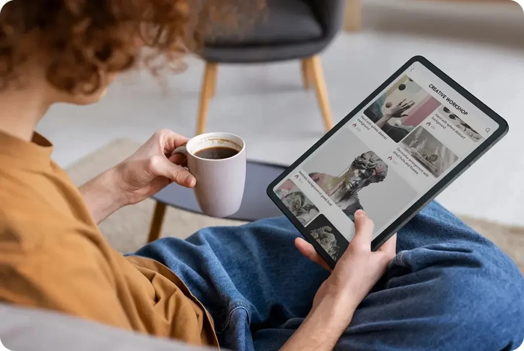 TCL Nxtpaper 11 Gen 2 to nowy i niedrogi tablet z Androidem. Osoba trzymająca tablet na kolanach przegląda kreatywne inspiracje, jednocześnie pijąc kawę z białego kubka, siedząc wygodnie w fotelu.