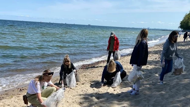 Grupa młodych osób zbiera śmieci na piaszczystej plaży nad morzem, każda z nich trzyma plastikowy worek, a w tle widać spokojną wodę oraz niewielką liczbę statków na horyzoncie.