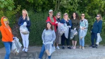 Grupa kobiet stoi na plaży, trzymając w rękach worki wypełnione śmieciami, ubrana w wygodne ubrania i rękawiczki ochronne, za nimi roślinność i drzewa, scena utrzymana w pogodnej atmosferze wspólnej działalności.