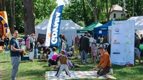 Rodziny z dziećmi biorą udział w festynie plenerowym, dzieci grają w gry na kocu, dorośli rozmawiają, wokół ustawione są namioty promocyjne oraz kolorowe baloniki, atmosfera jest radosna i rodzinna.