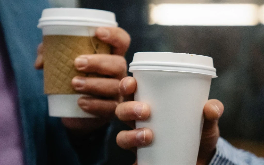 Kawa na wynos? Niby nic strasznego, ale wydatki rosną Dwie osoby trzymające kubki papierowe z gorącym napojem, jeden kubek okryty brązowym kartonowym rękawem, drugi biały, na tle nieostrego pomieszczenia.