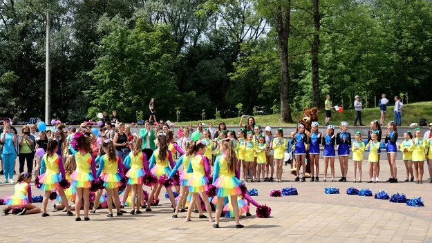 Kolorowo ubrana grupa dziewcząt wykonuje układ taneczny na placu, otoczona przez młodzież w żółto-niebieskich strojach sportowych, w tle zielone drzewa i kibicujący obserwatorzy.