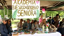 Grupa seniorów siedzi przy stole pod namiotem, na którym widnieje ręcznie wykonany napis 'Akademia Bieszczadzkiego Seniora'. Na stole rozłożone są prace rękodzielnicze, ozdoby i dekoracje wykonane przez uczestników. W tle widać innych ludzi spacerujący...
