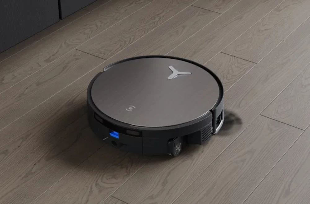 Robot sprzątający o okrągłym kształcie poruszający się po jasnej, drewnianej podłodze, wyposażony w czujniki i nowoczesny design.