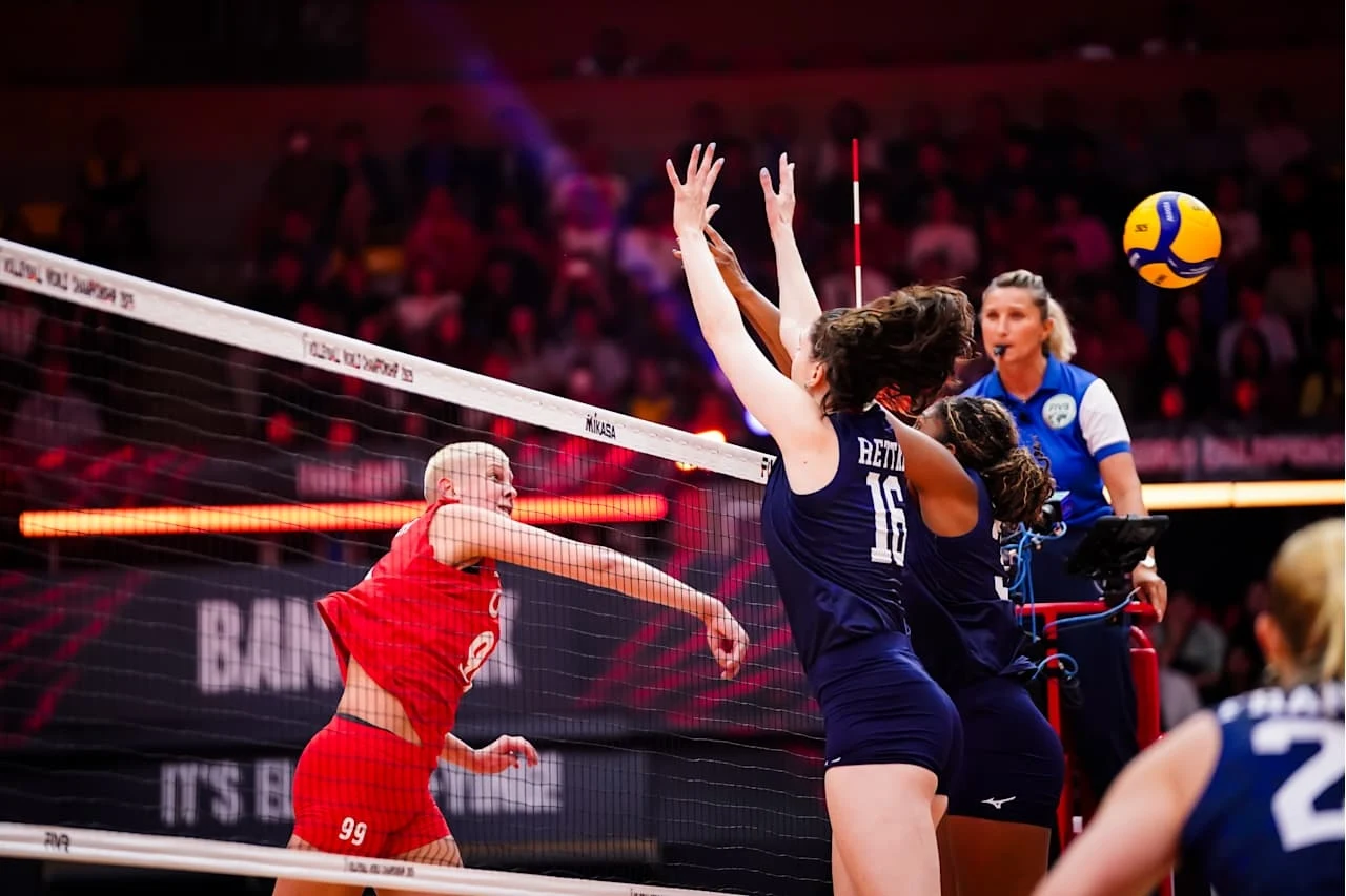 Kadr z meczu Turcja - USA, w ataku Ebrar Karakurt Zawodniczka w czerwonym stroju wykonuje atak podczas meczu siatkówki, naprzeciwko dwie przeciwniczki w niebieskich strojach próbują blokować piłkę nad siatką, a z boku obserwuje sędzia.