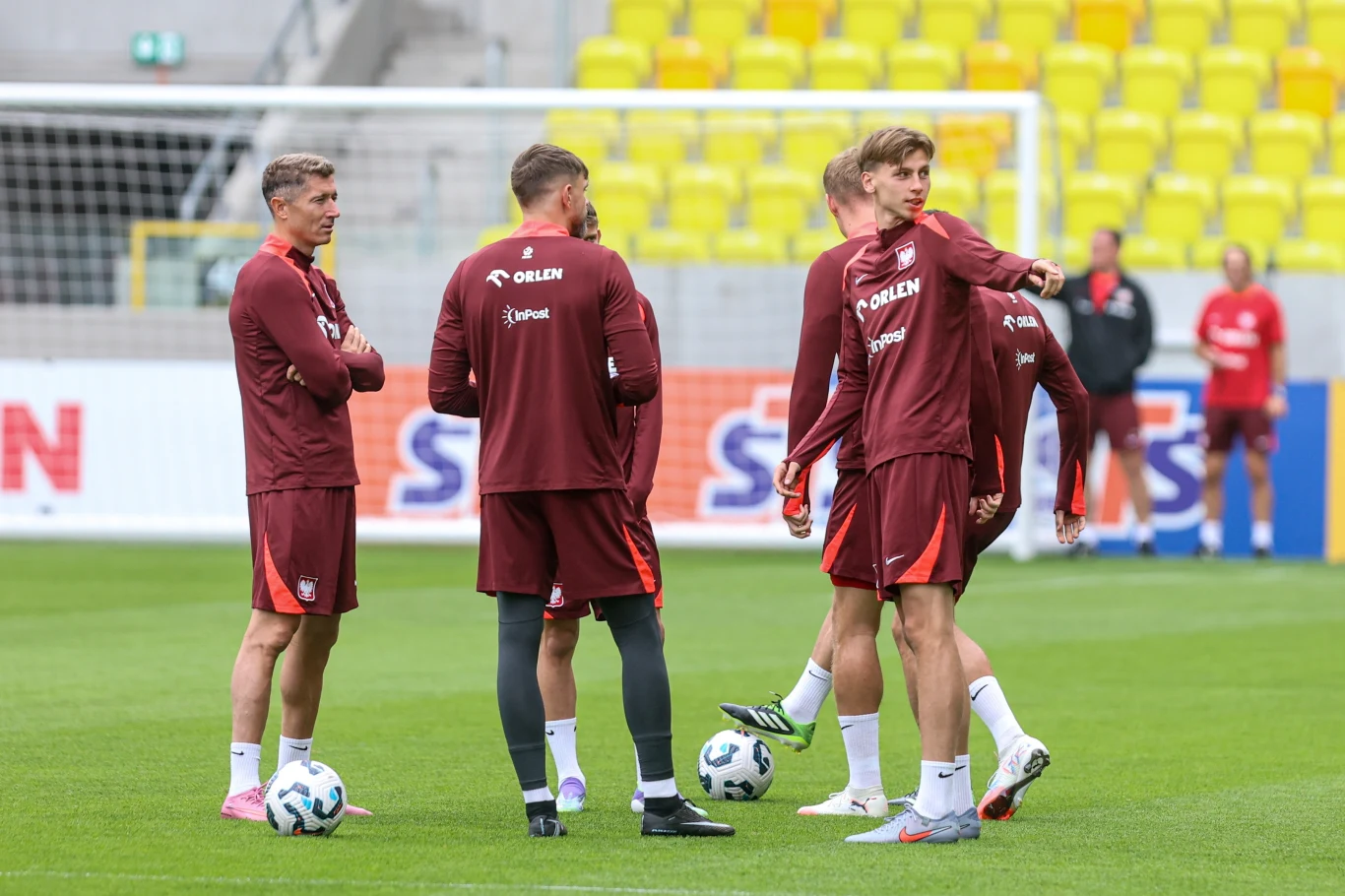 Grupa piłkarzy w strojach treningowych stoi na boisku, rozmawiając i przygotowując się do ćwiczeń, w tle bramka oraz żółte krzesełka stadionowe.