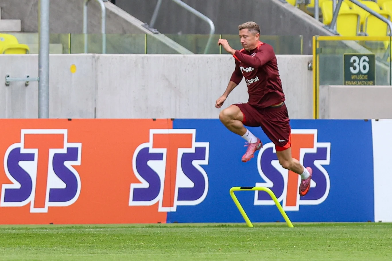 Piłkarz w bordowym stroju sportowym przeskakuje przez żółty płotek podczas treningu na stadionie piłkarskim, w tle widoczne są reklamy i żółte krzesełka na trybunach.