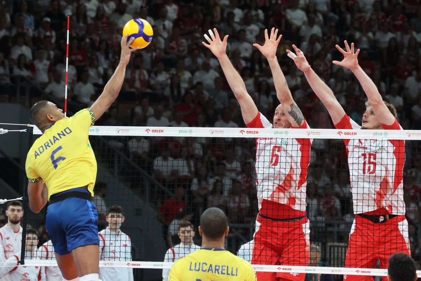 Bartosz Kurek i Jakub Kochanowski w bloku, a w ataku Adriano Moment rywalizacji w meczu siatkówki, zawodnik w żółtym stroju z drużyny Brazylii atakuje piłkę nad siatką, podczas gdy dwóch zawodników w biało-czerwonych strojach reprezentacji Polski próbuje zablokować atak, w tle pełne trybuny kibiców.