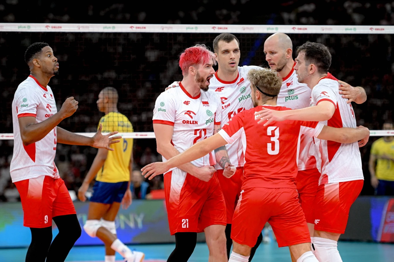 Siatkarze reprezentanci Polski w meczu z Brazylią Reprezentacja Polski w siatkówce mężczyzn ciesząca się po zdobyciu punktu podczas meczu, grupa zawodników ubranych w biało-czerwone stroje otacza się wzajemnie na boisku, w tle widoczny jest zarys przeciwnika w żółtym stroju oraz siatka.