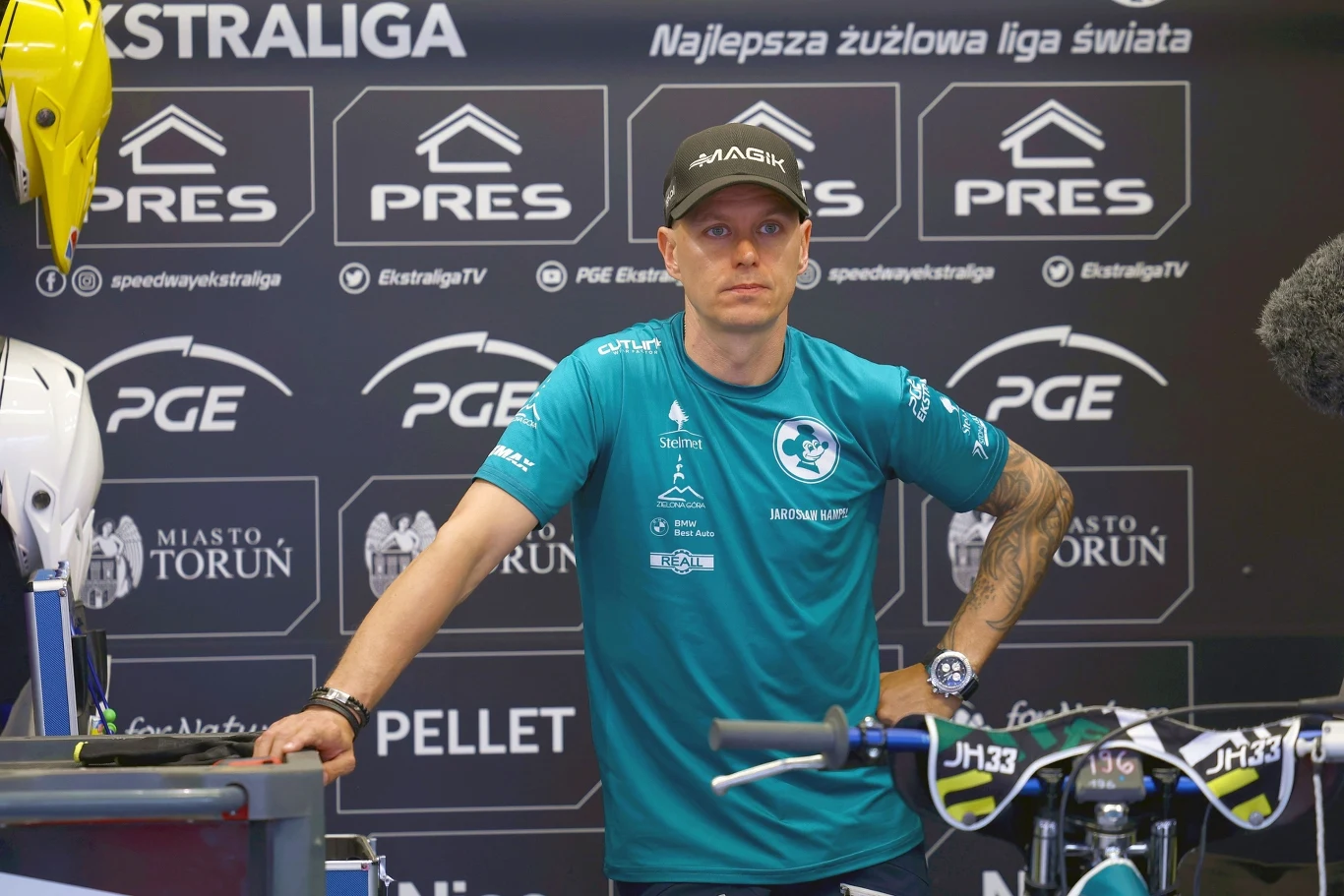 Mężczyzna ubrany w sportową koszulkę i czapkę z daszkiem stoi obok motocykla żużlowego na tle ścianki promocyjnej z logotypami sponsorów, na jednym z boków widoczne żółte kaski ochronne.