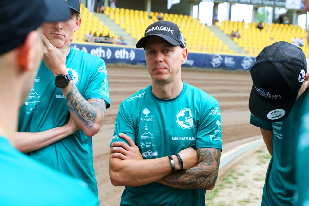 mężczyzna w sportowej koszulce i czapce z napisem MAGIK stoi z założonymi rękami na tle stadionu z żółtymi krzesełkami, obok niego dwóch innych mężczyzn w podobnych strojach