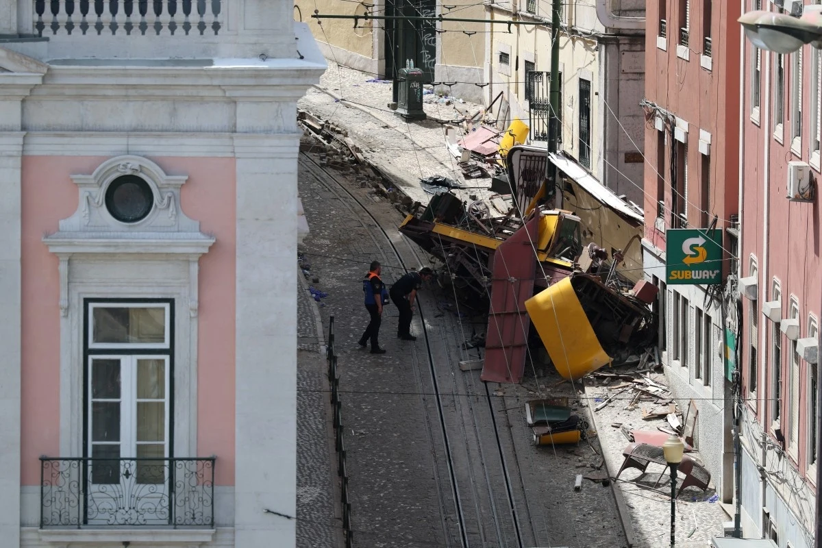 Lizbona. Wypadek kolejki Gloria. Wskazano, jakiej narodowości są ofiary Zawalony wagonik tramwaju częściowo rozbity na stromym, brukowanym zboczu, otoczony gruzem i fragmentami konstrukcji, po bokach stoją budynki mieszkalne oraz widoczny jest szyld restauracji.