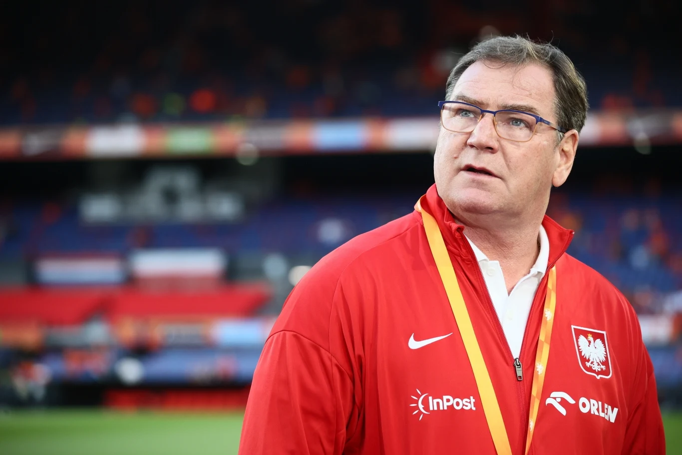 Jan Urban zadebiutował w roli selekcjonera Mężczyzna w czerwonym dresie z polskim godłem i napisem Orły, stojący na tle stadionu sportowego, z zawieszoną na szyi akredytacją.