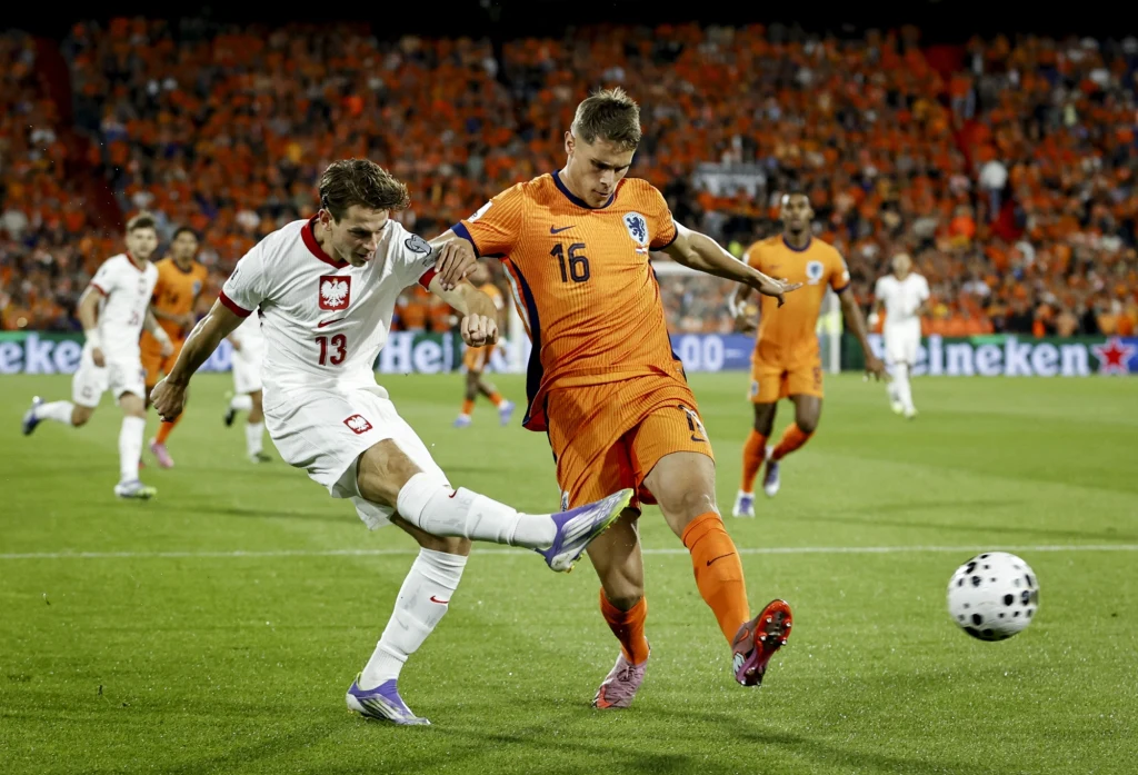 Jakub Kamiński w meczu Polska - Holandia Dwóch piłkarzy walczy o piłkę podczas meczu, zawodnik w białym stroju reprezentuje Polskę, a zawodnik w pomarańczowym stroju reprezentuje Holandię, w tle widać tłum kibiców na trybunach.