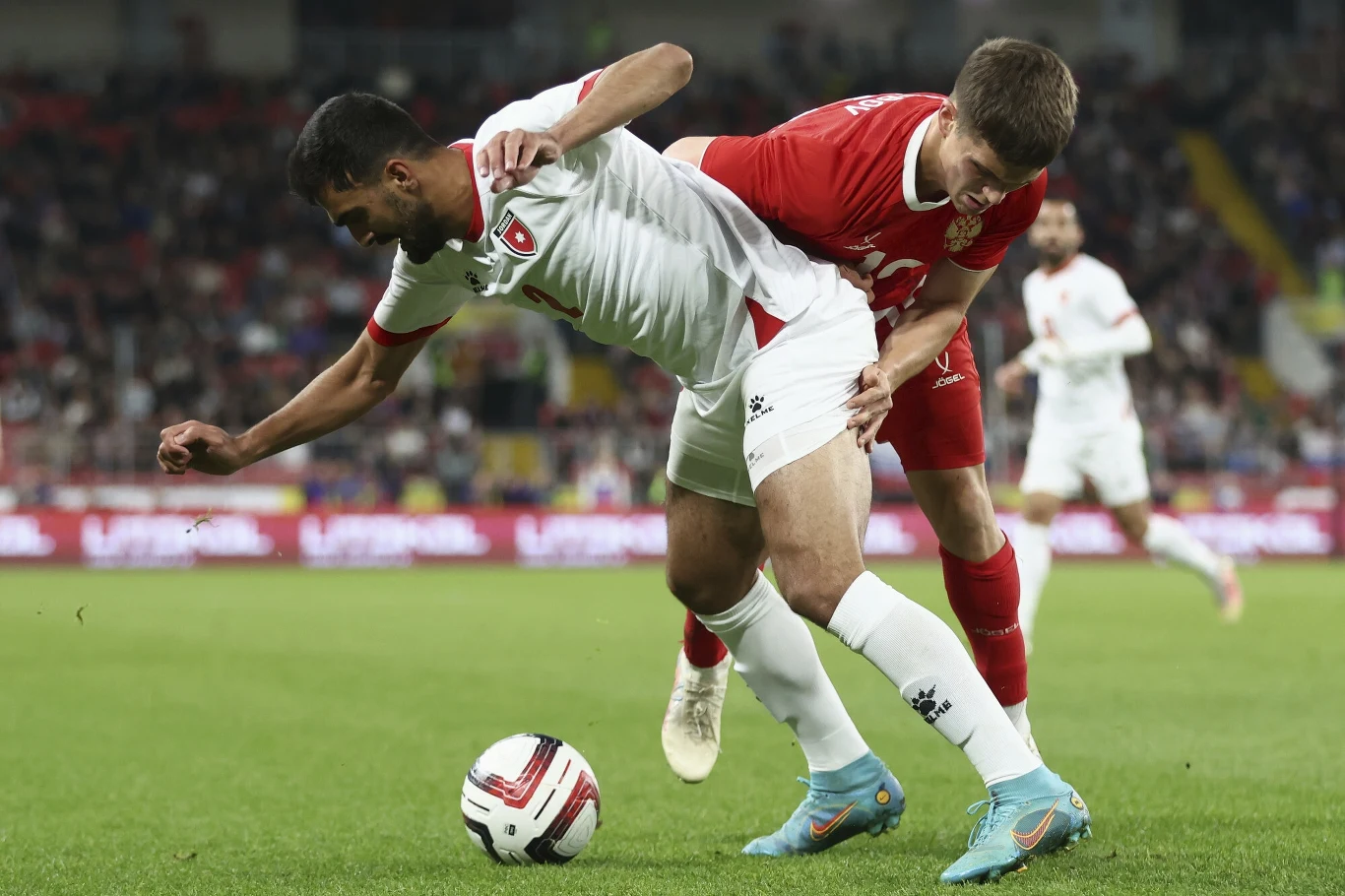 Dwóch piłkarzy walczących o piłkę na boisku podczas meczu, jeden ubrany w czerwony, drugi w biały strój, w tle rozmazane trybuny z kibicami