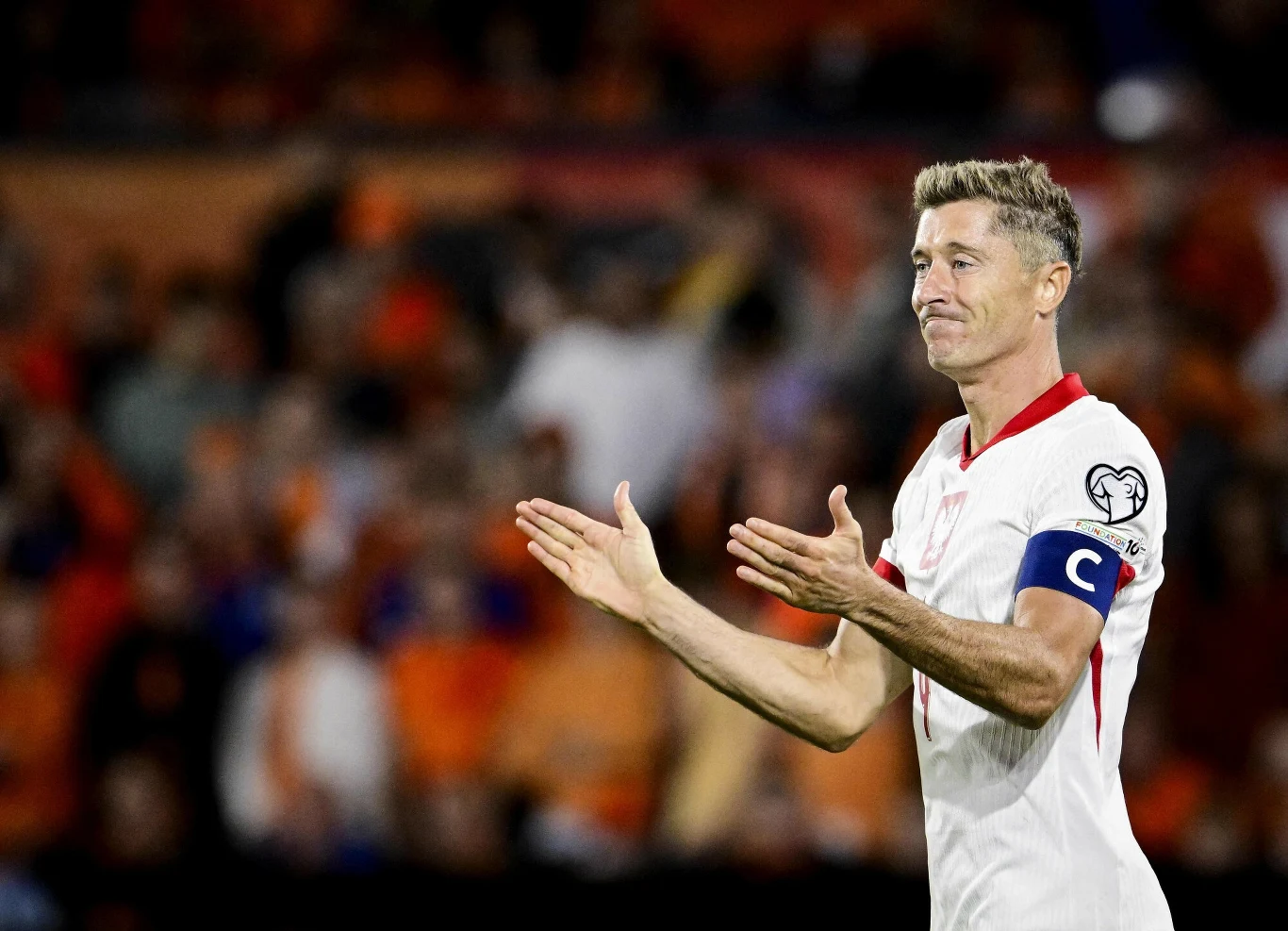 Robert Lewandowski w meczu z Holandią Piłkarz w białym stroju z opaską kapitana na ramieniu klaszcze na stadionie podczas meczu, w tle rozmyty tłum widzów.
