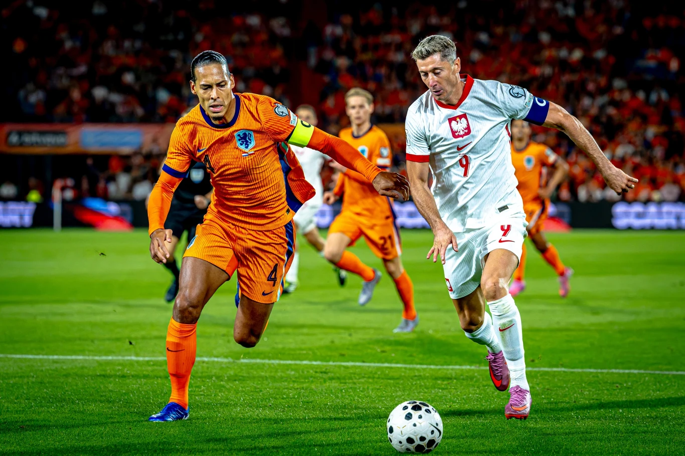 Robert Lewandowski w pojedynku z Virgilem van Dijkiem Dwóch piłkarzy rywalizuje o piłkę podczas meczu – jeden w pomarańczowym stroju reprezentacji Holandii, drugi w białym stroju reprezentacji Polski; w tle widoczny fragment boiska oraz inni zawodnicy.