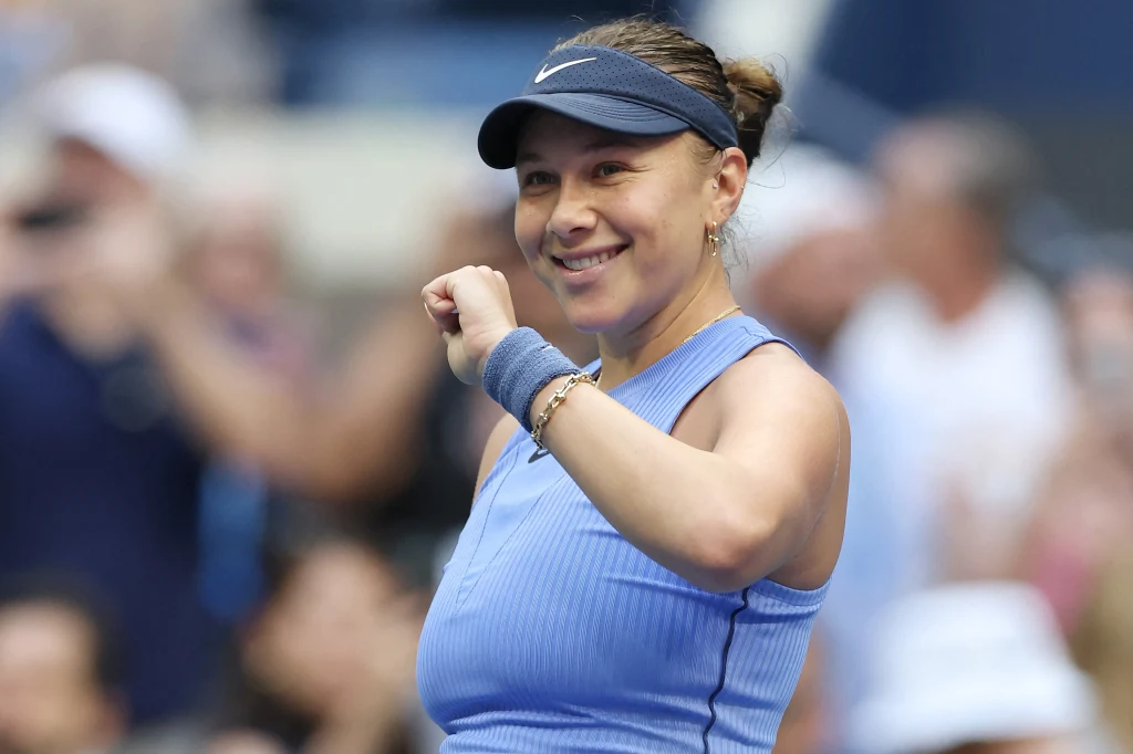 Uśmiechnięta kobieta w sportowej niebieskiej koszulce, z opaską na głowie i opaską na nadgarstku, gestykuluje zaciśniętą pięścią na tle rozmytej publiczności podczas wydarzenia sportowego.