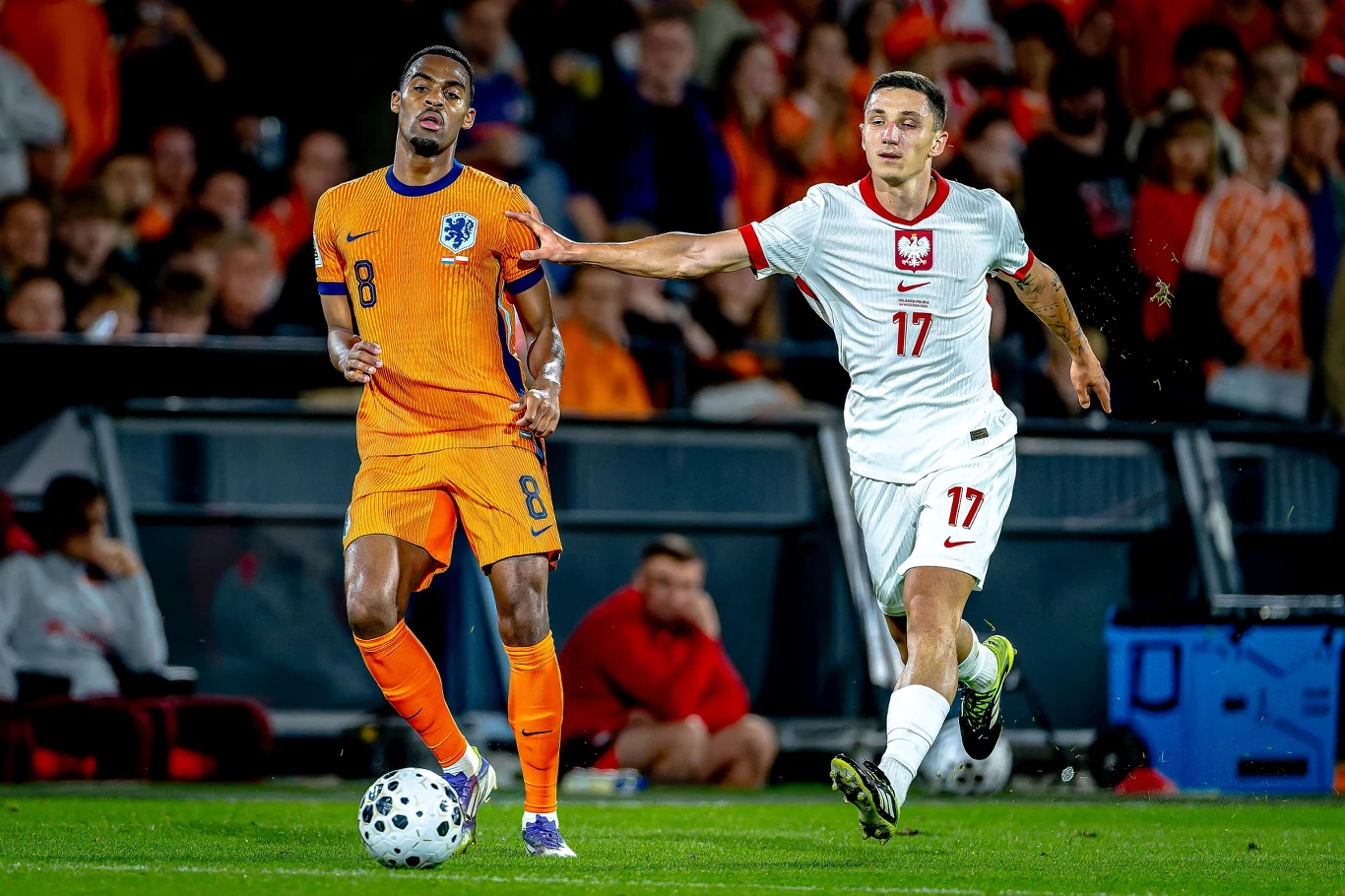 Ryan Gravenberch i Bartosz Slisz Dwóch piłkarzy podczas meczu piłki nożnej, jeden w pomarańczowej, drugi w białej koszulce, między nimi trwa walka o piłkę, w tle kibice na trybunach.