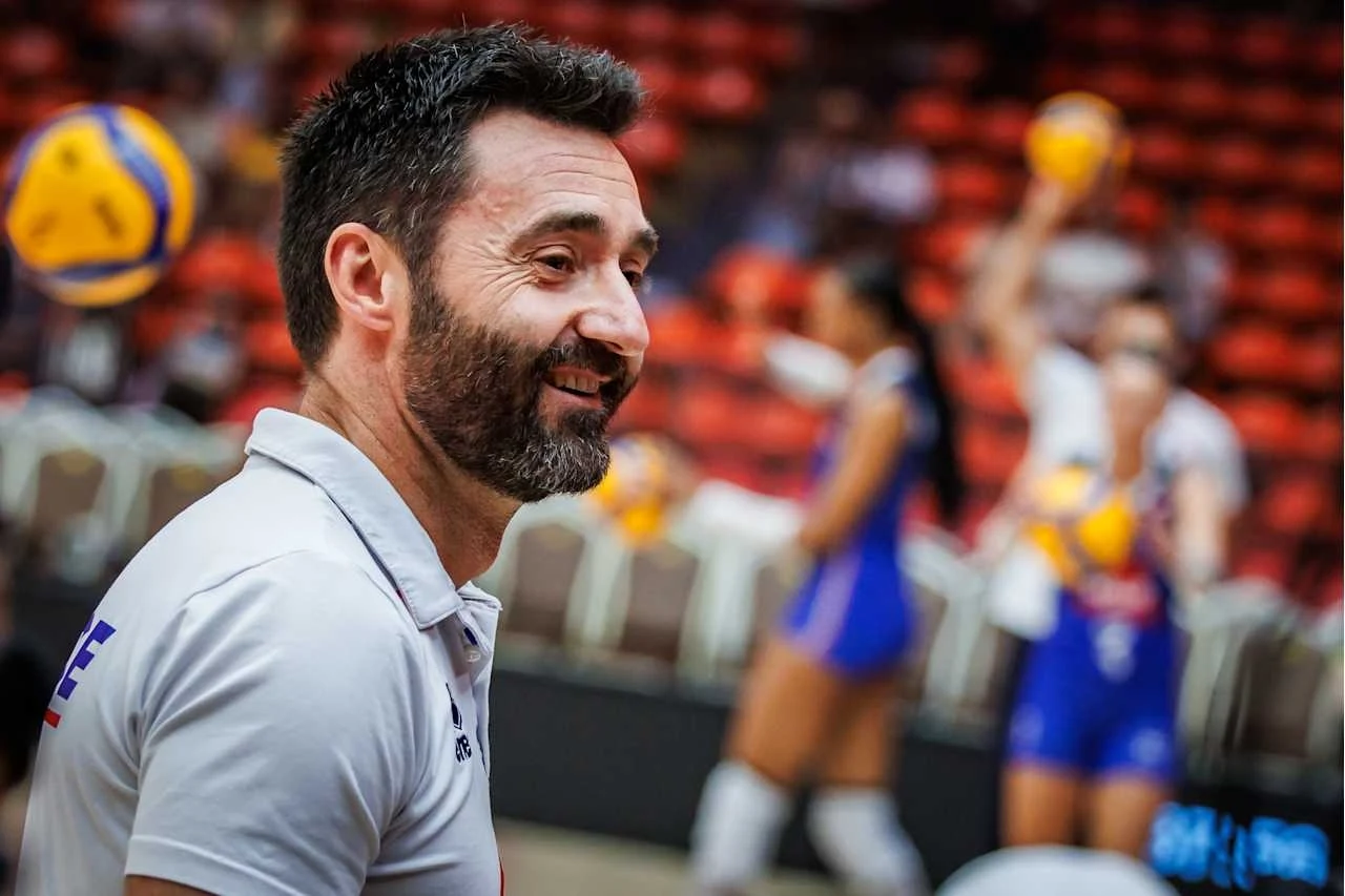 Trener uśmiechnięty na tle hali sportowej, w której siatkarki w niebieskich strojach przygotowują się do gry z piłkami w rękach.