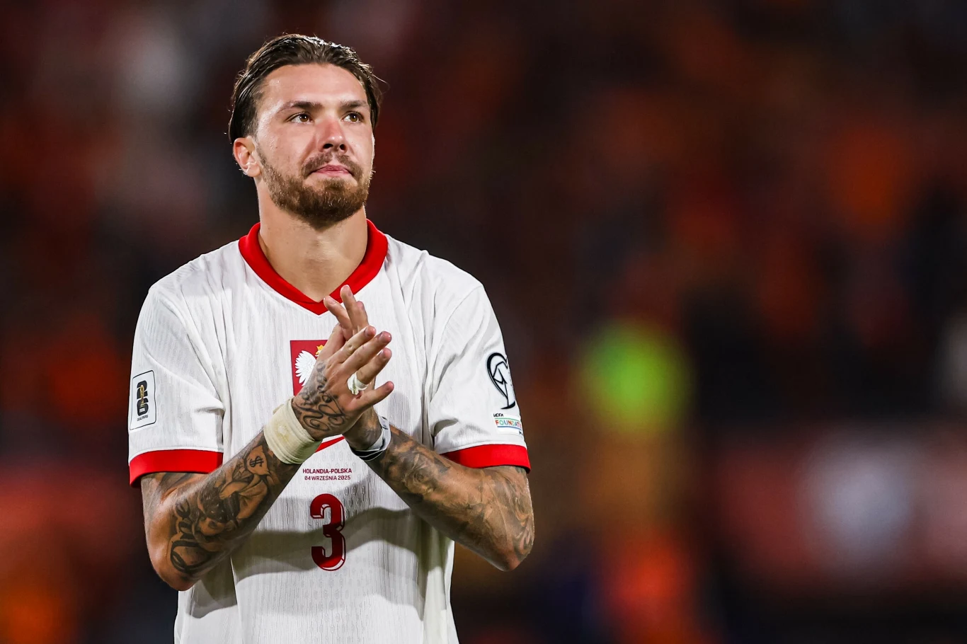 Przemysław Wiśniewski w meczu z Holandią, 04.09.2025 r. Piłkarz w białej koszulce z numerem 3 i narodowym herbem Polski stoi na boisku, klaszcząc w dłonie, w tle zamazana publiczność.