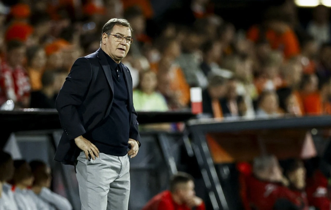 Mężczyzna w marynarce i okularach stoi na tle rozmytego tłumu na stadionie sportowym, obserwując wydarzenia na boisku, w otoczeniu osób ubranych w sportowe stroje.