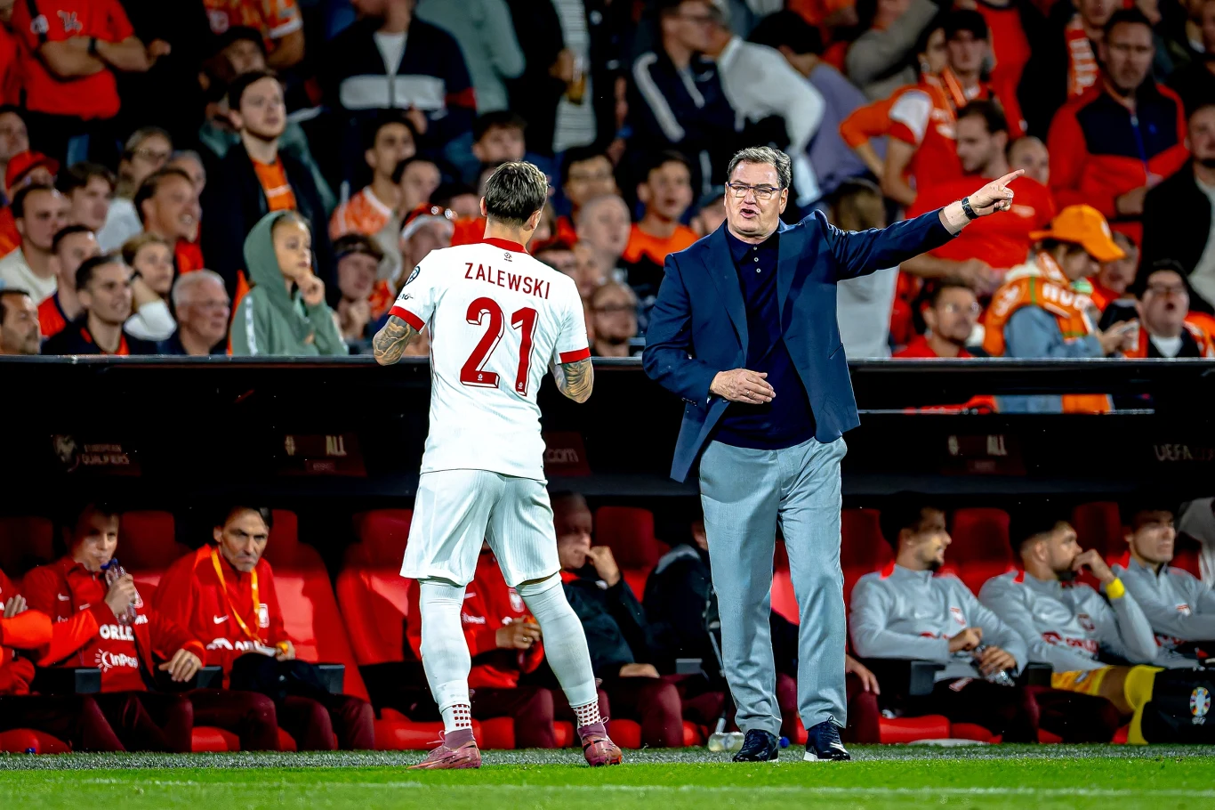 Piłkarz w białym stroju z numerem 21 i napisem ZALEWSKI rozmawia z trenerem na linii bocznej boiska, wokół trybuny wypełnione kibicami ubranym głównie w czerwone barwy.