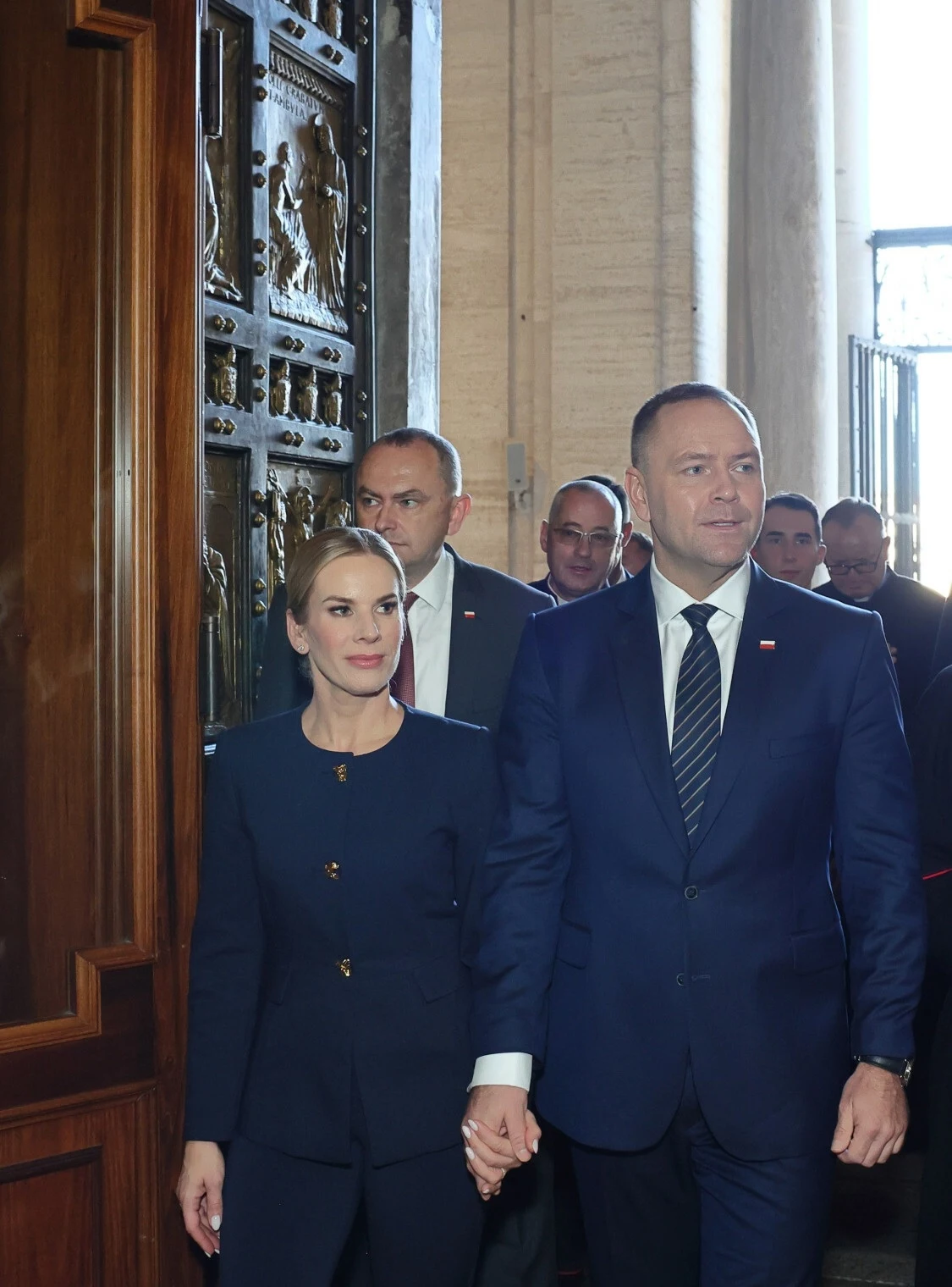 Marta Nawrocka kolejny raz pokazała, że ceni sobie w modzie minimalizm i prostotę Para ubrana formalnie w garnitury i ubrania wizytowe wchodzi do wnętrza przez duże dekoracyjne drzwi, za nimi idzie grupa mężczyzn również w strojach oficjalnych.