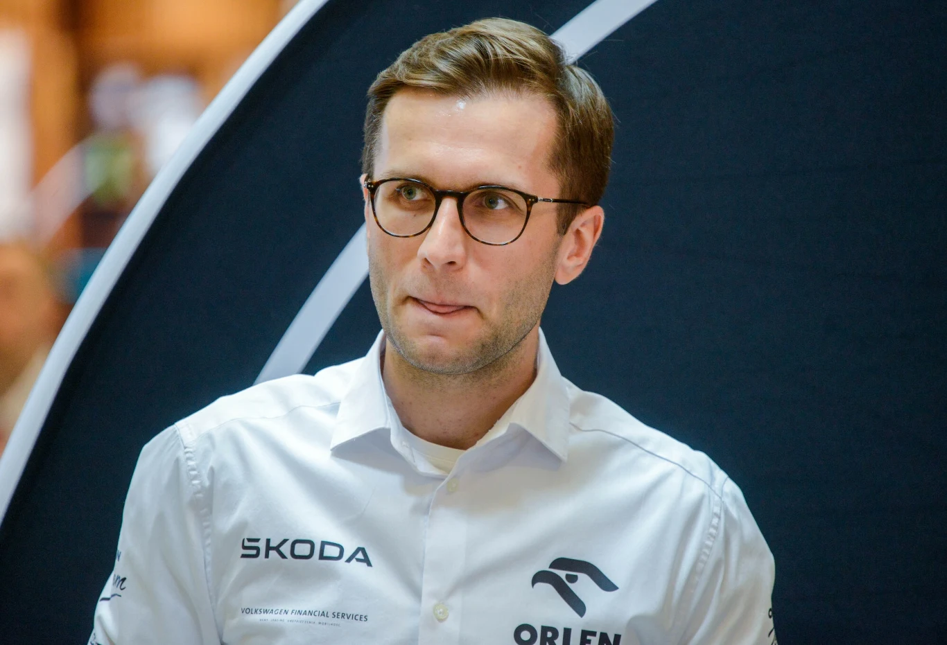 Mikołaj Marczyk Młody mężczyzna w okularach, ubrany w białą koszulę z logotypami firm Orlen i Skoda, stoi na tle nowoczesnego wnętrza, patrzy lekko w bok z lekko wysuniętym językiem.