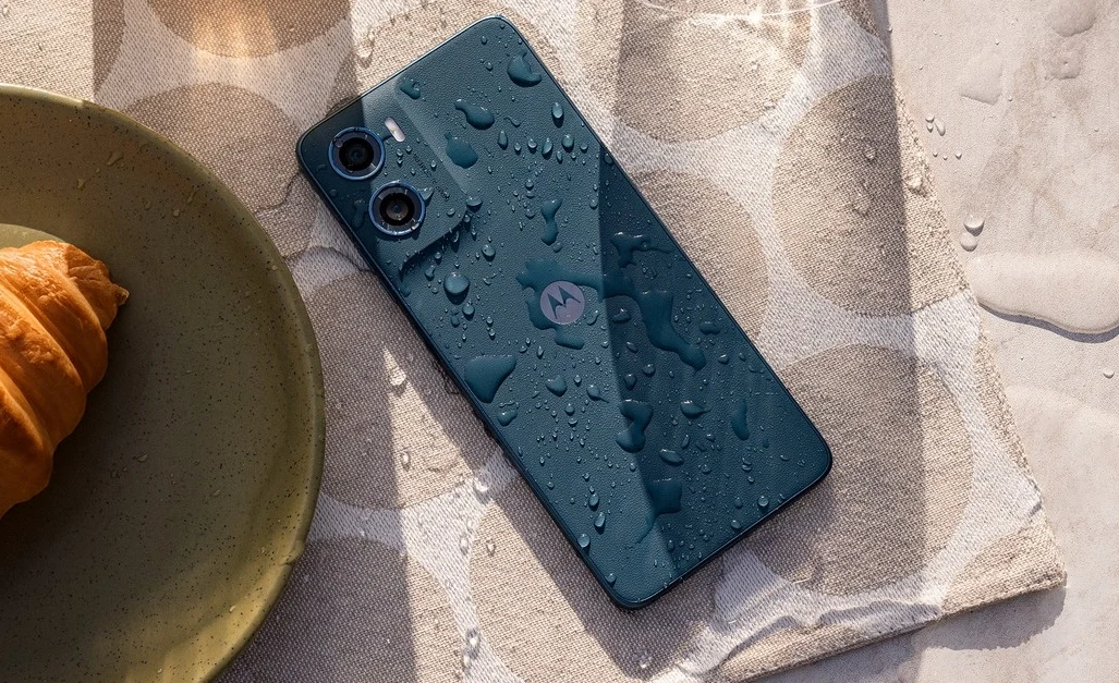 Nowoczesny smartfon z podwójnym aparatem leżący na lnianej serwetce, pokryty kroplami wody, obok znajduje się talerz z croissantem.