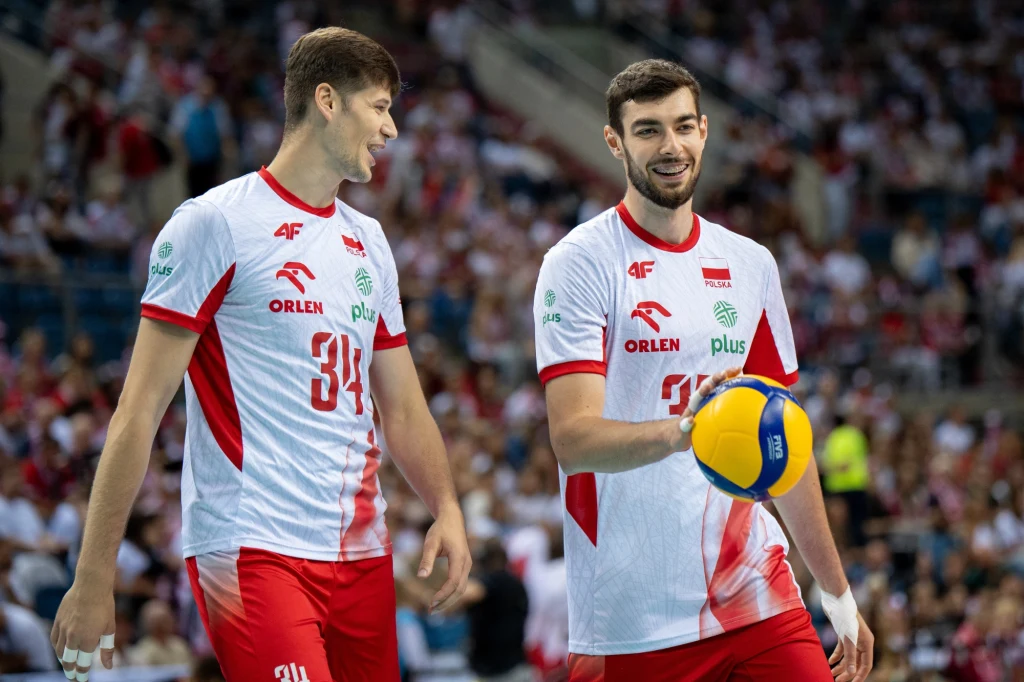 Dwóch siatkarzy w białych koszulkach z czerwonymi elementami stoi na boisku, jeden z nich trzyma piłkę do siatkówki, obaj uśmiechają się, w tle widoczna rozmyta publiczność.