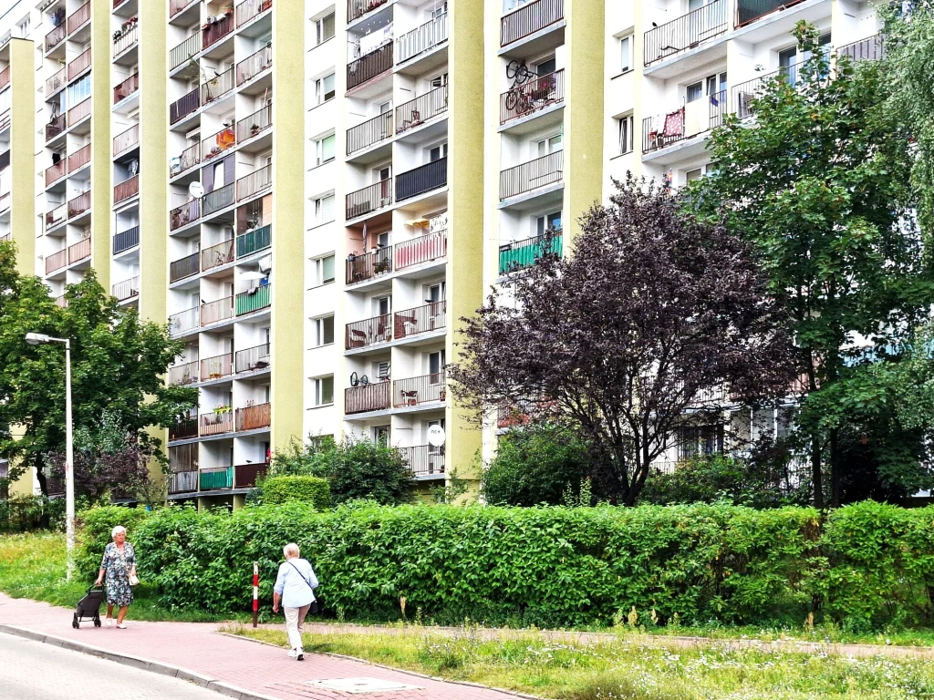 Wysoki blok mieszkalny z licznymi balkonami otoczony zielenią i drzewami, przed budynkiem chodnik, po którym idą dwie starsze kobiety, jedna z nich ciągnie wózek na kółkach.