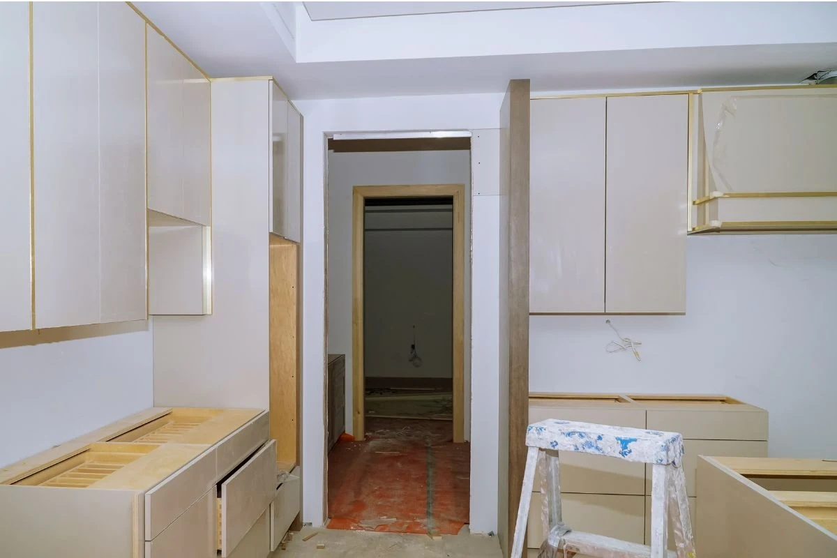 Jasna kuchnia w trakcie remontu, na podłodze folia ochronna, szafki bez frontów, drabina malarska, jasne ściany i nowoczesne zabudowy meblowe.