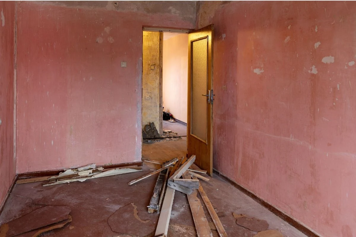 Puste pomieszczenie o różowych, zniszczonych ścianach z widocznymi śladami zużycia, otwarte drzwi prowadzą do drugiego, także zaniedbanego pokoju, na podłodze leżą rozrzucone elementy drewniane i resztki materiałów budowlanych.